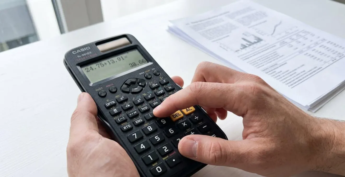 Gros plan de mains utilisant une calculatrice moderne posée sur un bureau blanc épuré, avec un document bancaire complètement flouté en arrière-plan, éclairé par une lumière latérale naturelle créant un effet bokeh