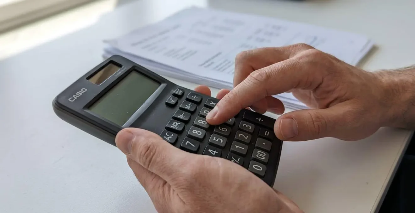 Gros plan de mains utilisant une calculatrice moderne posée sur un bureau blanc épuré, avec un document bancaire complètement flouté en arrière-plan, éclairé par une lumière latérale naturelle créant un effet bokeh